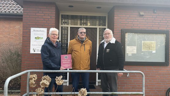 Drei Männer vor einem Haus. Der Mann ganz links hält ein Buch in die Kamera