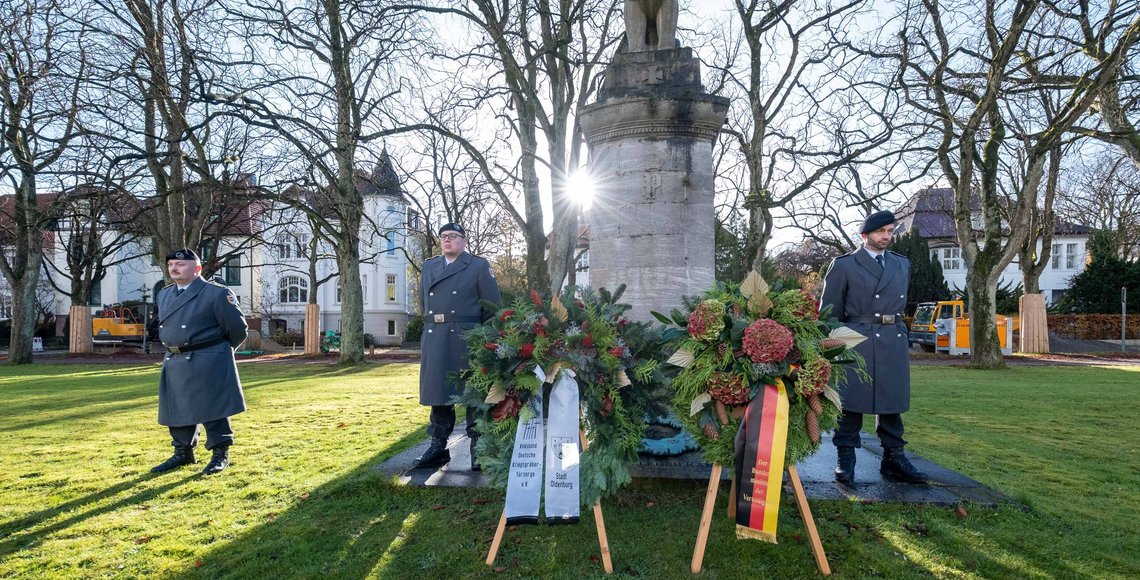 Drei Soldaten stehen neben einem Kriegerdenkmal, vor dem Ehrenmal stehen zwei Kränze