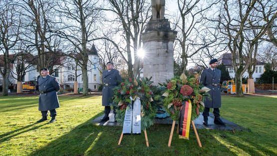 Drei Soldaten stehen neben einem Kriegerdenkmal, vor dem Ehrenmal stehen zwei Kränze