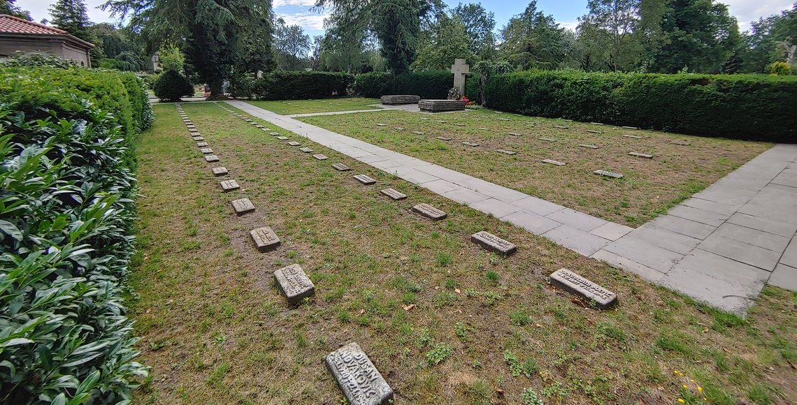 Zu sehen ist die Kriegsgräberstätte Osnabrück-Eversburg. Man sieht die einzelnen Grabsteine gereiht. Im Hintergrund sieht man Bäume und Gebüsch.