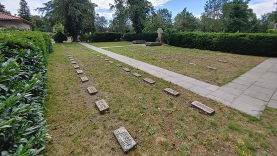 Zu sehen ist die Kriegsgräberstätte Osnabrück-Eversburg. Man sieht die einzelnen Grabsteine gereiht. Im Hintergrund sieht man Bäume und Gebüsch.