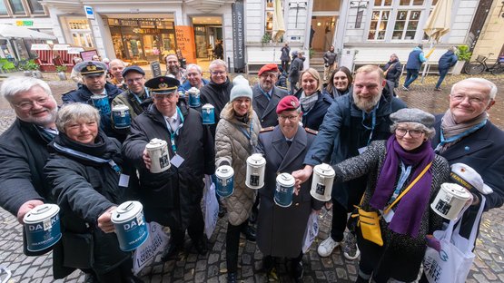 Menschengruppe blickt in Richtung Kamera und hält Sammeldose des Volksbunds ausgestreckt vor sich 