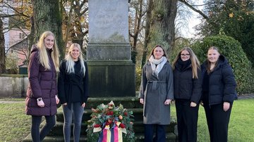 Zu sehen sind die Auszubildenden. Jeweils links und rechts stehen sie neben einem Denkmal und einem Gedenkkranz. Sie schauen alle lächelnd in die Kamera.
