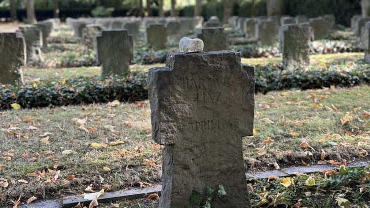 Auf dem Foto sieht man mehrere Kriegsgräber. Die Steinform ist ein Kreuz. Ein Grab ist im Vordergrund, die weiteren Gräber im Hintergrund sind verschwommen zu erkennen.